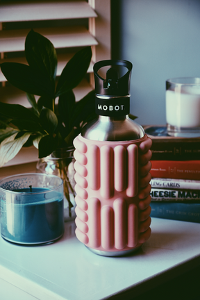 blush pink 40oz big bertha Mobot foam roller water bottle on a table next to a candle plant and books